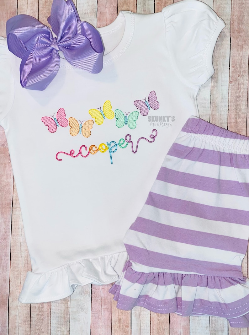 May include: A white baby outfit with a rainbow butterfly design and the name "cooper" embroidered on the front. The outfit includes a short-sleeved shirt with a ruffled hem and a pair of striped shorts with a ruffled hem.