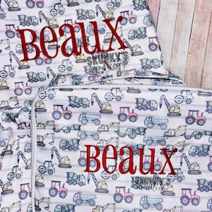 May include: A matching set of a backpack and lunch bag. Both are white with a pattern of construction vehicles in various colors and a red embroidered name. The background has gray and white stripes.