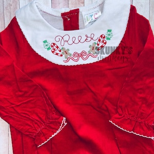 May include: Red corduroy baby dress with a white Peter Pan collar and embroidered Christmas details, including candy canes, gingerbread men, and the name "Reese".