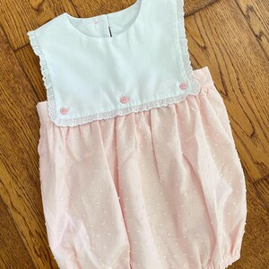 May include: A baby's bubble romper in white and pink. The top is white with a rounded collar and pink buttons. The bottom is pink with a textured pattern. The romper has a lace trim.