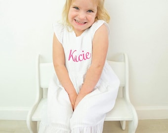 May include: A young girl wearing a white dress with a pink bow in her hair. The dress has the name "Kacie" embroidered on the front in pink thread. The girl is sitting on a white wooden chair. The image is captioned "Skunky's Monkeys".