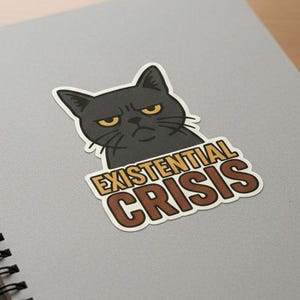 May include: A sticker featuring a grumpy black cat with yellow eyes. The sticker has the words "EXISTENTIAL CRISIS" in brown and gold lettering. The sticker is on a gray spiral notebook.