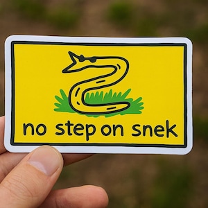 Puede incluir: Una pegatina amarilla con un contorno negro presenta una serpiente enroscada sobre hierba verde. El texto "no step on snek" está debajo de la serpiente en negro. La pegatina se sostiene sobre un fondo borroso verde y marrón.