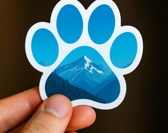 Puede incluir: Adhesivo blanco con forma de pata con un diseño de paisaje de montaña azul. Las almohadillas de la pata son de color azul claro. La escena de la montaña presenta un pico nevado, árboles y tonos de azul.