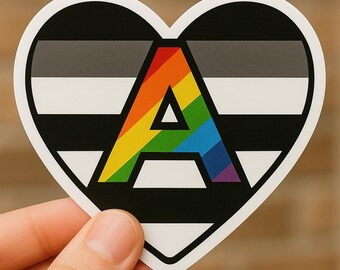 May include: A heart-shaped sticker featuring the letter "A" in rainbow colors, set against a black and white striped background. The sticker has a white border and is held up against a brick wall.