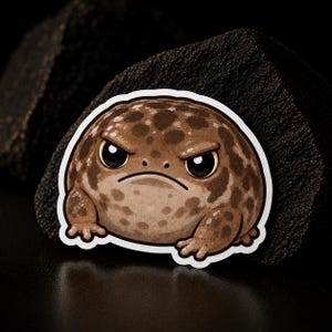 May include: A die-cut sticker featuring a round, brown toad with dark brown spots and a grumpy facial expression. The toad has large black eyes, a downturned mouth, and small front feet, rendered in a flat, illustrated graphic style.