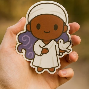 May include: A sticker featuring a cartoon character with brown skin, wearing a white robe and head covering. The character has purple hair and is holding a white dove. The sticker is held in a hand.