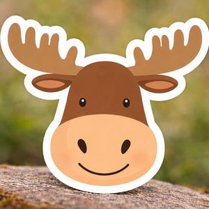 Könnte beinhalten: Aufkleber mit einem Cartoon-Elch mit weißem Rand. Der Elch hat braune Hörner, einen braunen Kopf und ein hellbraunes Gesicht mit einem lächelnden Mund und schwarzen Augen. Der Hintergrund ist verschwommen grün und braun.