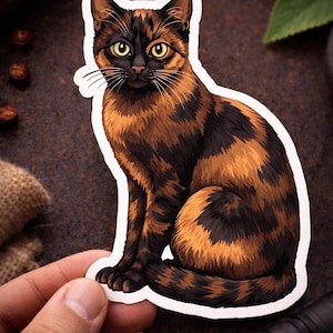 May include: A sticker featuring a sitting tortoiseshell cat with a brown and black patterned coat. The cat has yellow eyes and a white outline. The sticker is held by a hand.