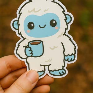 May include: A die-cut sticker featuring a cartoon yeti holding a blue mug with a brown beverage. The yeti has white fur, a light blue face, and rosy cheeks. The sticker is held by a hand against a blurred green background.