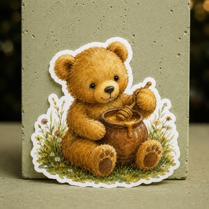 Peut inclure: Un autocollant découpé représentant un ours en peluche brun assis dans un champ de petites fleurs sauvages jaunes et blanches. L'ours tient une cuillère à miel en bois dans un pot en céramique brun rempli de miel doré, avec des coussinets visibles.