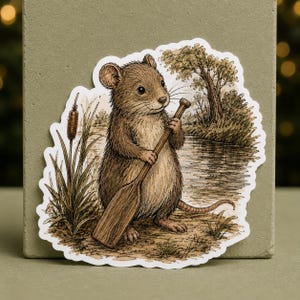May include: A die-cut sticker featuring a brown mouse standing on a riverbank, holding a wooden paddle. The illustration includes a cattail plant, a flowing river, and a tree in the background, rendered in a detailed, sketch-like artistic style.