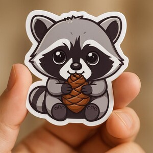 May include: A cute cartoon raccoon sticker with a gray and black color scheme. The raccoon is holding a brown pinecone. The sticker has a white border and is set against a blurred background.