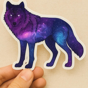 Puede incluir: Una pegatina con un diseño de lobo con un patrón de galaxia. El lobo está representado en tonos de morado, azul y blanco, con un efecto de galaxia arremolinada en su cuerpo. La pegatina tiene un borde blanco y está sujeta por una mano.