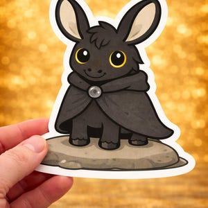May include: A sticker of a cartoon donkey in a black cape with a silver clasp, standing on a rock. The donkey has large eyes with yellow highlights and long ears. The background is a blurred gold colour.