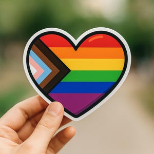 May include: A heart-shaped sticker with a rainbow design and a white border. The rainbow is divided into six horizontal stripes, with a progressive pride flag on the left side. The sticker is held by a hand.