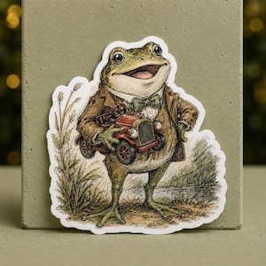 Op de afbeelding: Een gestanste sticker met een antropomorfe kikker in een bruin jasje en een groene vlinderdas. De kikker houdt een kleine rode vintage speelgoedauto vast. De illustratie bevat hoog gras en een vijver op de achtergrond in aardetinten.