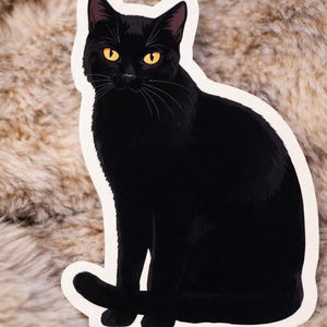 May include: A black cat sticker with a white border. The cat is sitting with its tail curled and has bright yellow eyes. The sticker is on a textured, light brown surface.