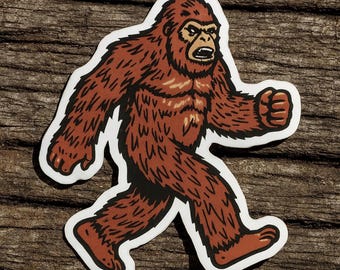 May include: A sticker featuring a cartoon illustration of Bigfoot walking. The creature is brown with a white outline and black accents. The image is set against a wooden background.