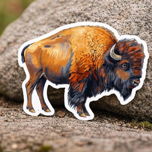Op de afbeelding: Een sticker met een gedetailleerde illustratie van een bizon. De bizon is afgebeeld in tinten oranje, bruin en zwart, met een witte omtrek. De sticker is geplaatst op een getextureerd rotsachtig oppervlak.
