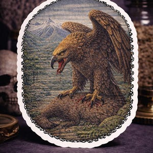 May include: Oval sticker with a detailed illustration of a brown eagle perched on a brown animal, set against a mountain landscape. The eagle's wings are spread, beak open, with blood visible. The sticker has a decorative border.
