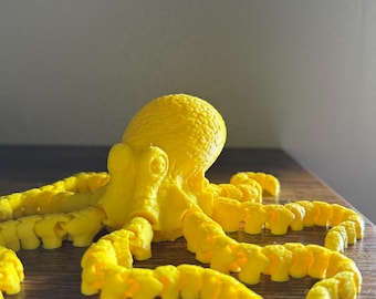 May include: A bright yellow 3D printed octopus with eight tentacles. The octopus is sitting on a wooden surface.