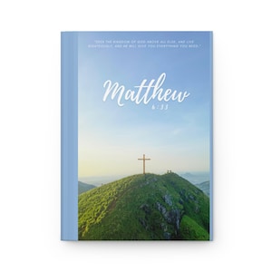 May include: A blue and white book cover with the text "Seek the kingdom of God above all else, and live righteously, and he will give you everything you need." and "Matthew 6:33". The cover features a photo of a wooden cross on a green hilltop with a blue sky in the background.