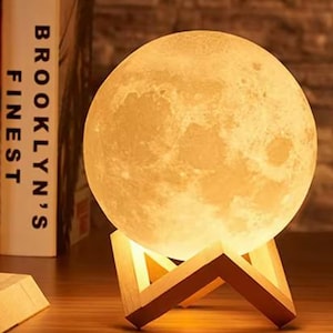 Puede incluir: Una lámpara blanca en forma de luna con una textura realista de superficie lunar. La lámpara está sobre un soporte de madera con un diseño geométrico.