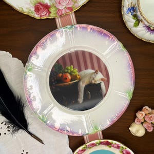 May include: A decorative plate with a pearlescent rim featuring a central image of a white cat resting near a still life of fruit. The plate is surrounded by other vintage china and a black feather.