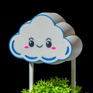 May include: A white cloud-shaped decoration with a smiling face, blue outline, and pink cheeks. The cloud sits atop a white stand above a bed of green artificial plants, all against a black background.