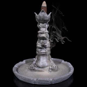 May include: A gray incense burner featuring a stack of frog figurines, topped with a lotus flower design. A lit incense cone sits atop the lotus, with wisps of smoke rising against a black background. The burner sits in a round, shallow dish.