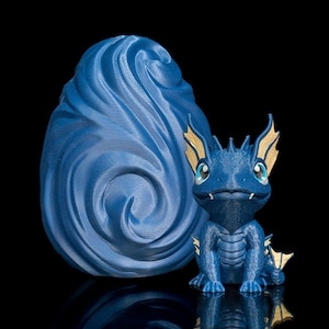 May include: A blue dragon figurine with gold accents and a matching blue egg-shaped sculpture. The dragon has large eyes and a friendly expression. The egg has a swirling design. The objects are set against a black background.