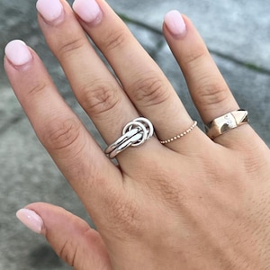 Può includere: Una mano che mostra quattro anelli. Gli anelli includono un anello a nodo argentato, un anello dorato con perline e un anello dorato con tre piccoli diamanti. Le unghie sono dipinte con smalto rosa chiaro.