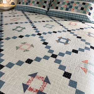 May include: A white quilt with a geometric pattern of blue, black, and red squares and arrow-shaped designs. Two matching pillowcases with a patchwork design and a blue plaid border rest on the bed. The quilt is a traditional, handmade item.