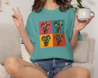 May include: Teal t-shirt featuring a pop art style design of four potted plants on a colorful background. The plants have large, patterned leaves. The shirt is made of a soft material and is suitable for casual wear.