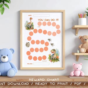 Op de afbeelding: Een ingelijste beloningskaart met een licht houten frame. De kaart heeft een dinosaurus thema met illustraties en de tekst "YOU CAN DO IT!". De kaart bevat ruimtes voor een naam, start en beloning. De onderkant van de kaart luidt "REWARD CHART".