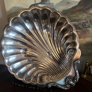 May include: A silver-colored, shell-shaped serving dish. The dish has a fluted design radiating from the center, with a decorative, ornate handle. The dish is likely made of metal and reflects light, creating a shiny appearance. The dish is sitting on a dark surface.