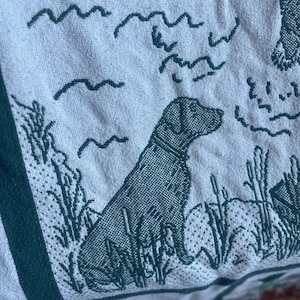 Pu&ograve; includere: Primo piano di una coperta tessuta con un design verde e bianco. La coperta raffigura un cane Labrador Retriever seduto in un campo di erba alta. Linee ondulate e altri elementi naturali sono anch'essi tessuti nel design.