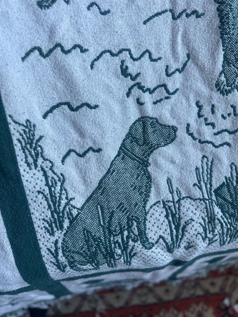 Pu&ograve; includere: Primo piano di una coperta tessuta con un design verde e bianco. La coperta raffigura un cane Labrador Retriever seduto in un campo di erba alta. Linee ondulate e altri elementi naturali sono anch'essi tessuti nel design.