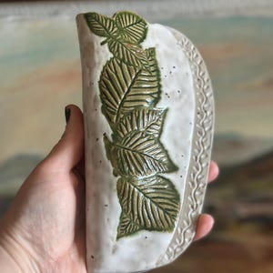 May include: A semi-circular ceramic wall vase with a textured off-white glaze and a vertical row of embossed green leaves. The curved edge features a delicate, repeating vine pattern, and the surface has subtle speckles throughout the finish.