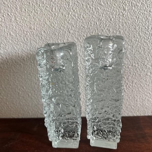 May include: Two clear glass candle holders with a textured, rough surface. The candle holders are rectangular with a square base and a hollowed-out top for candles. They are displayed on a dark wooden surface.