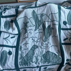 Pu&ograve; includere: Una coperta color crema con un bordo verde e illustrazioni di anatre e cani. La coperta presenta un motivo ripetuto di anatre in volo, un cane e piante acquatiche. La coperta ha un bordo a frange.