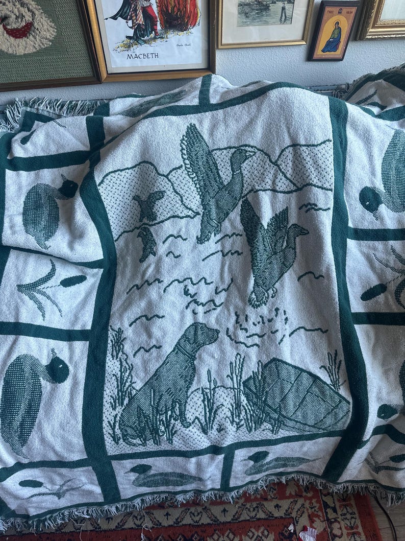 Pu&ograve; includere: Una coperta color crema con un bordo verde e illustrazioni di anatre e cani. La coperta presenta un motivo ripetuto di anatre in volo, un cane e piante acquatiche. La coperta ha un bordo a frange.