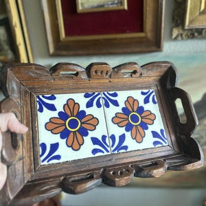 May include: A rectangular wooden tray with carved edges and handles. The tray's centre features two square tiles with a floral design in orange, blue, and yellow against a white background. The tray is likely used for serving or display.