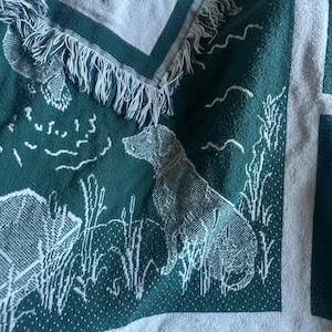 Pu&ograve; includere: Una coperta tessuta verde e bianca con un motivo di cane da caccia e scena all'aperto. La coperta presenta un bordo con frange e un motivo ripetuto di cani, anatre e canne. La coperta &egrave; realizzata in un materiale morbido e tessuto.