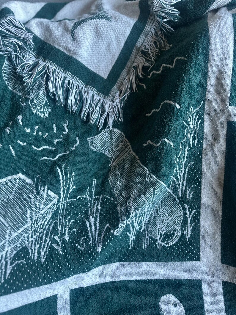 Pu&ograve; includere: Una coperta tessuta verde e bianca con un motivo di cane da caccia e scena all'aperto. La coperta presenta un bordo con frange e un motivo ripetuto di cani, anatre e canne. La coperta &egrave; realizzata in un materiale morbido e tessuto.