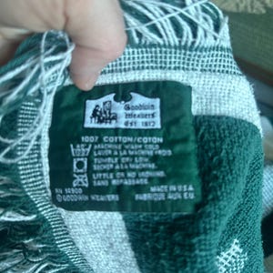 Pu&ograve; includere: Una coperta tessuta verde e bianca con un bordo a frange. La coperta ha un'etichetta che recita "Goodwin Weavers Est 1872" e "100% Cotone". L'etichetta include anche le istruzioni di lavaggio.