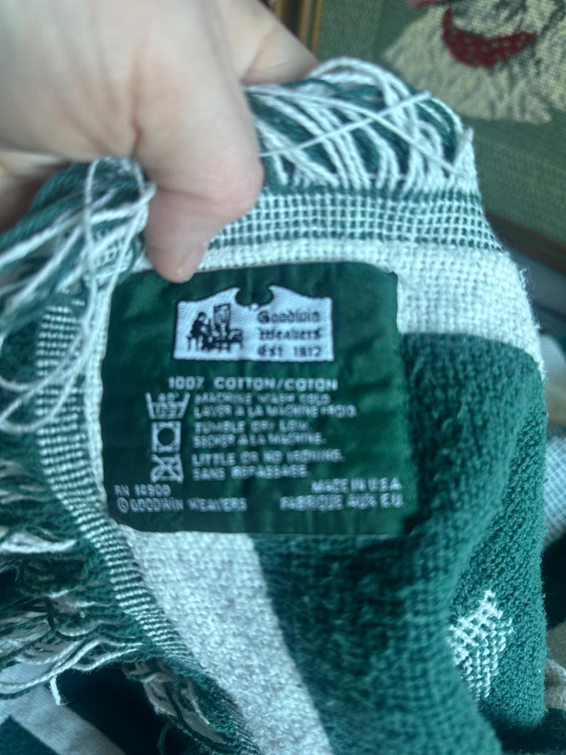 Pu&ograve; includere: Una coperta tessuta verde e bianca con un bordo a frange. La coperta ha un'etichetta che recita "Goodwin Weavers Est 1872" e "100% Cotone". L'etichetta include anche le istruzioni di lavaggio.