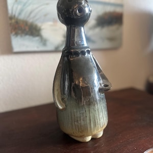 May include: A ceramic figurine featuring a tall, slender neck and a rounded head with an open mouth. The upper portion is finished in a glossy dark brown glaze, while the lower body transitions into a textured, muted green and beige base.