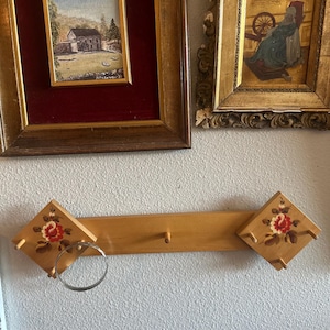 May include: A light-colored wooden wall rack featuring two diamond-shaped ends, each decorated with a hand-painted red and yellow floral motif. The rack includes three small wooden pegs and a circular metal ring attached to the left side.